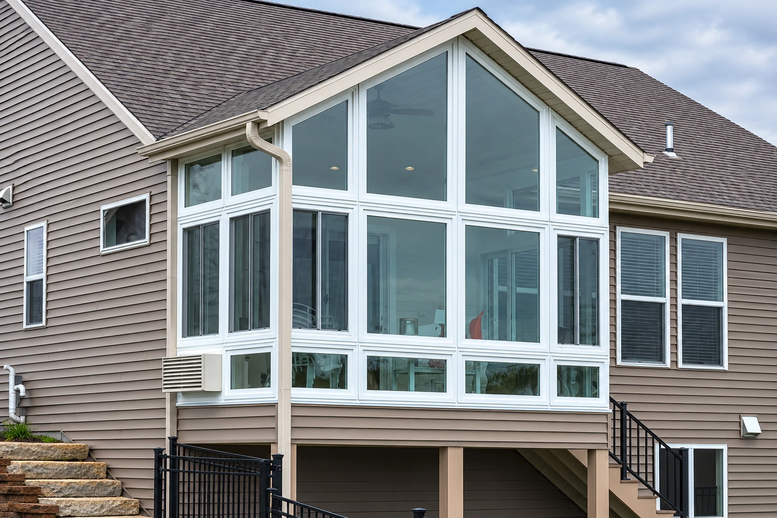 A custom sunroom with tall white-framed windows and a peaked glass upper section projects from a raised, gray-sided house.
