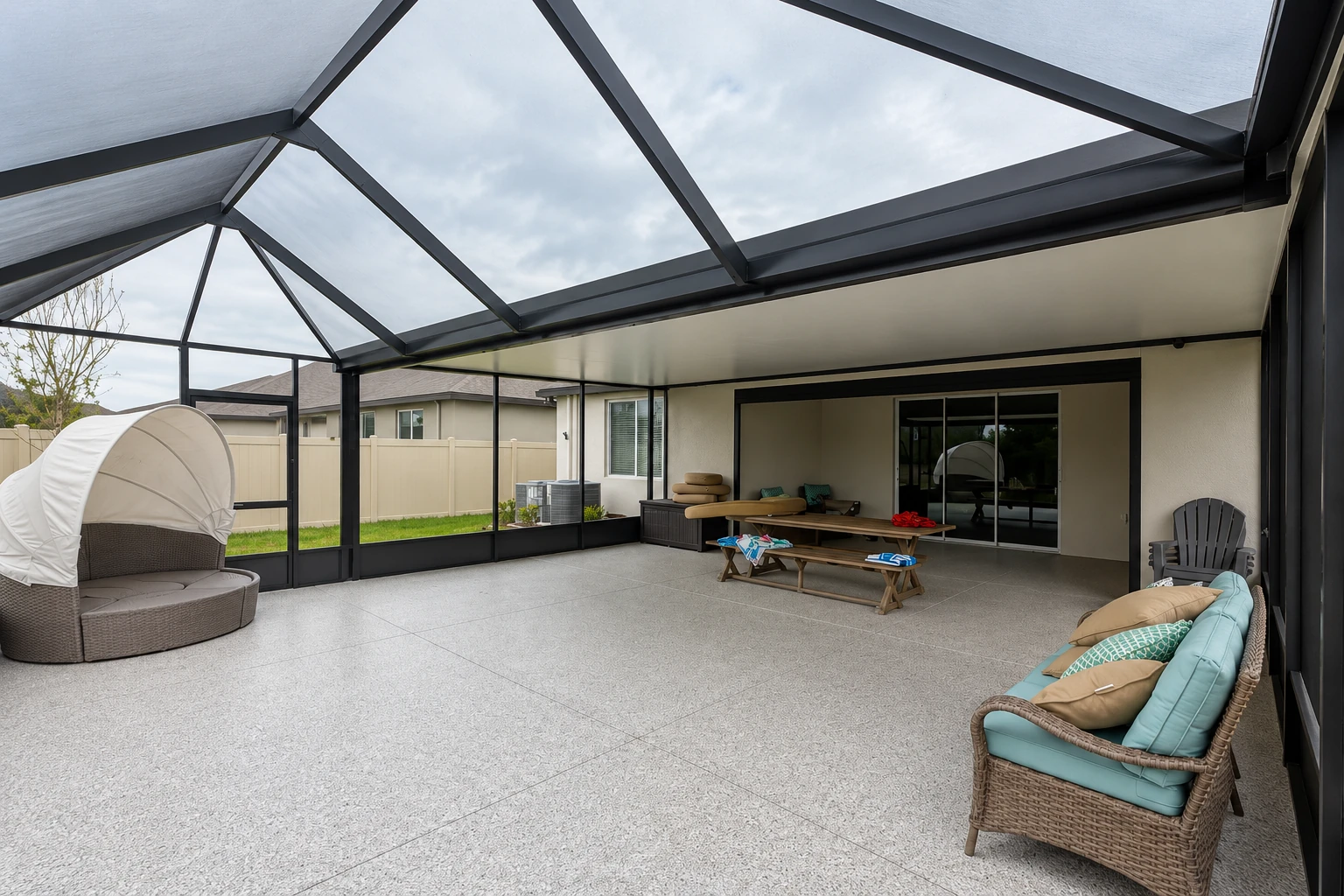 A spacious enclosed patio has a dark-framed glass roof and screened walls, with wicker seating and a picnic table.