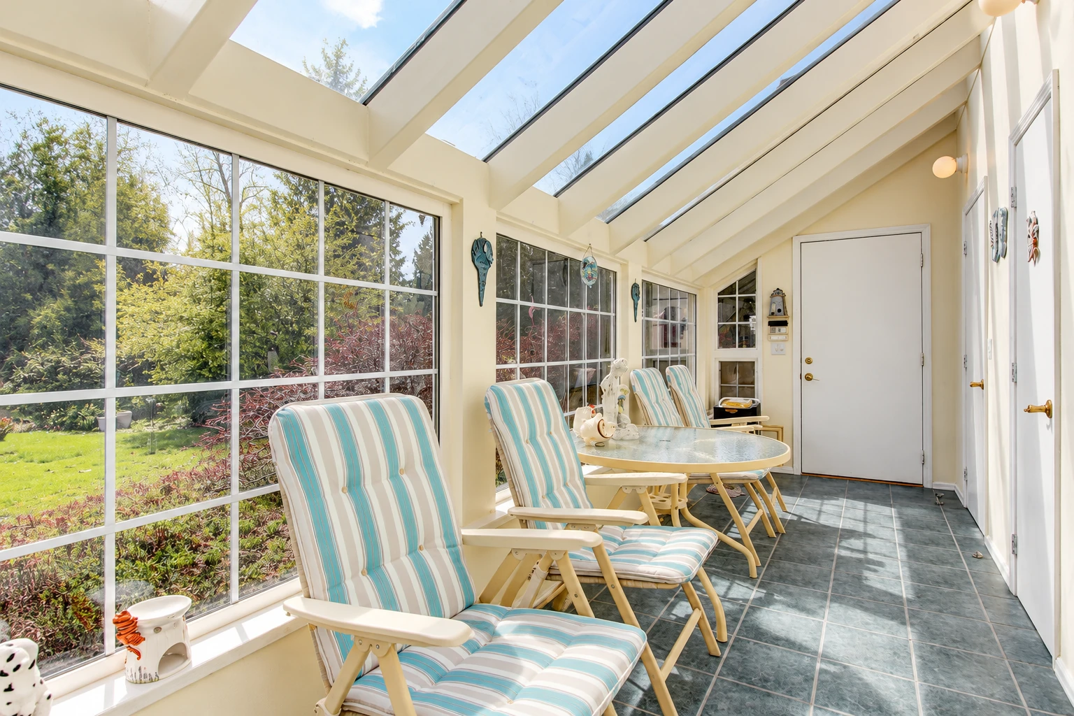 A bright four-season sunroom has a glass roof, tiled floor, striped chairs and a table beside large windows overlooking a garden.