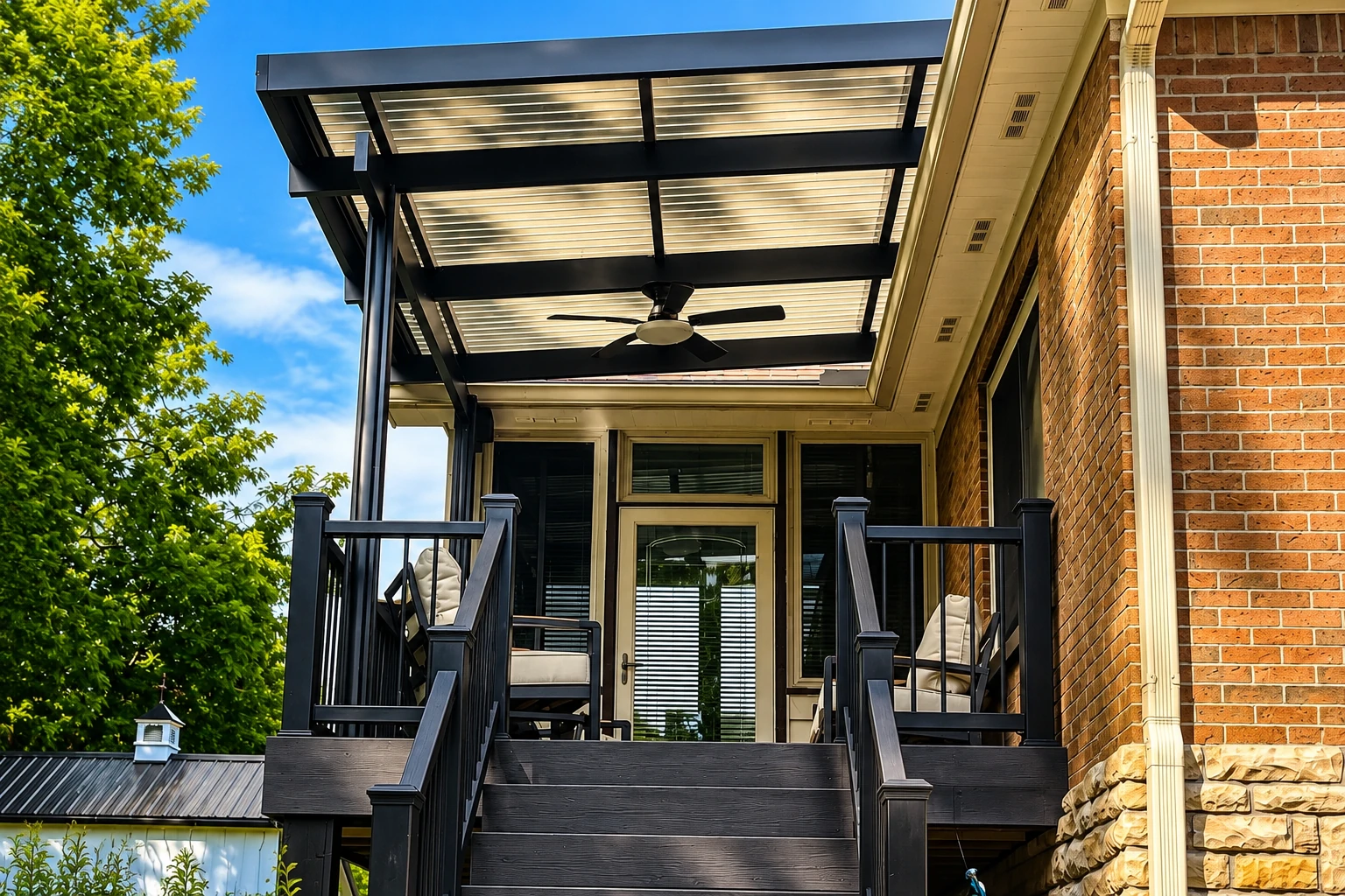 A black patio cover with translucent roof panels shades a raised deck beside a brick house.