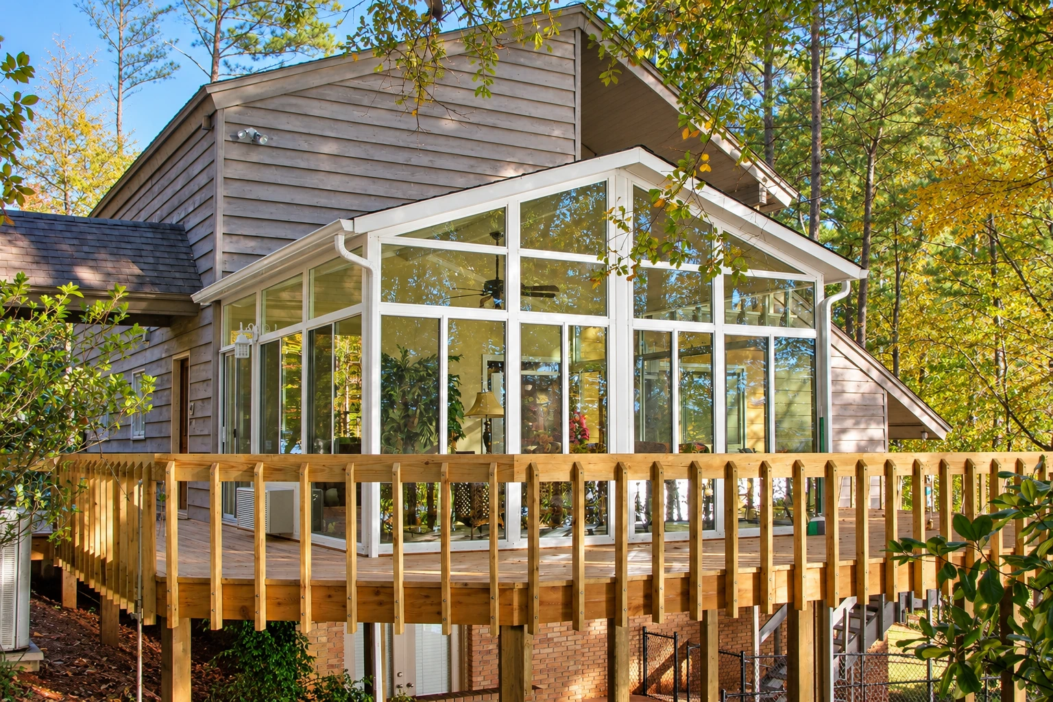 A white-framed glass sunroom projects from a wood-sided house, with a raised deck and railing among autumn trees.